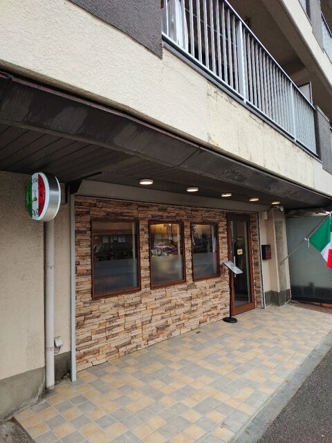 お店の外観。石調タイルの壁と丸いトリコローレの看板、店先にイタリア国旗